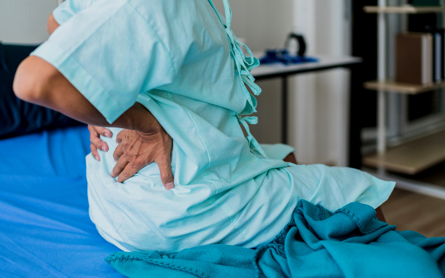 A man in a hospital gown holding his low back in pain.